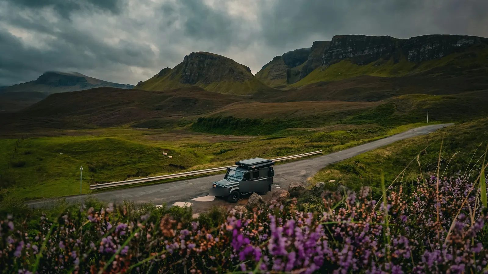 A 4x4 with a roof tent on a single-track road below green hills and cliffs, with purple heather in front